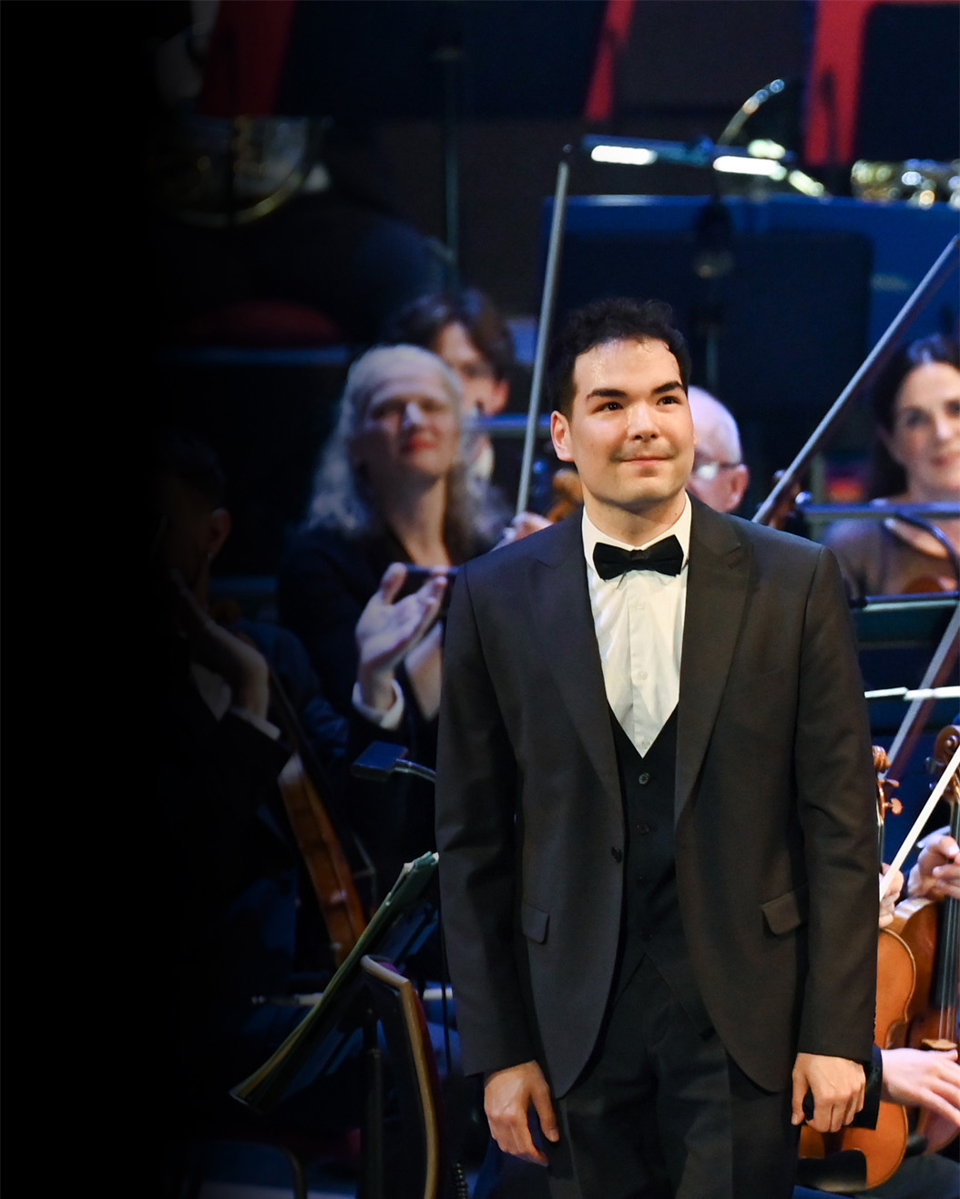 A man wearing smart attire, standing in front of an orchestra, also wearing smart attire and holding musical instruments, with the conductor clapping. 
